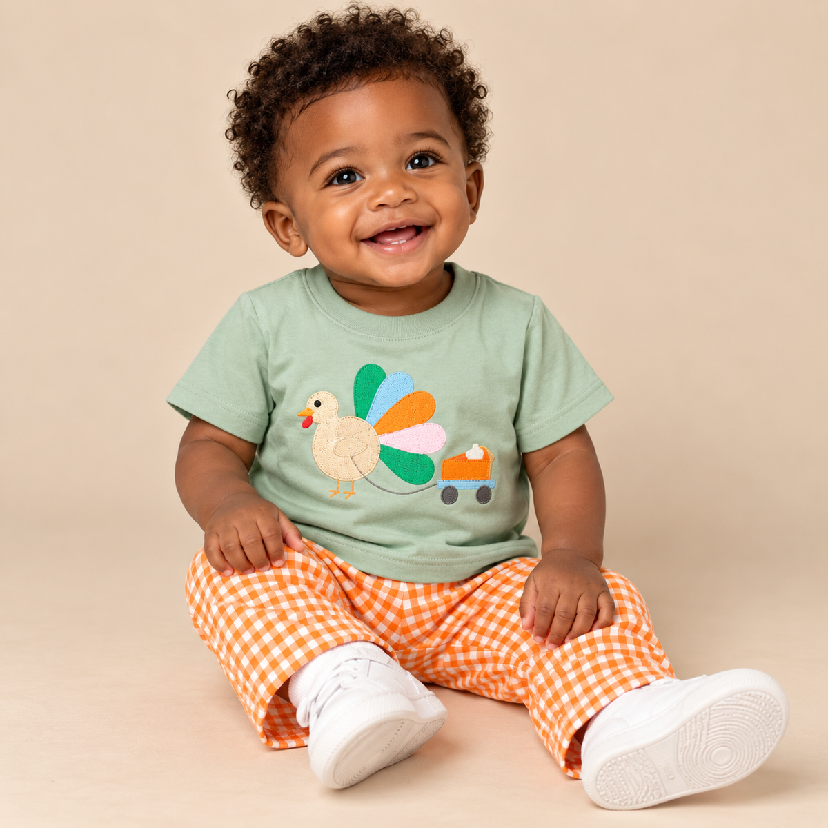 Infant boy wearing a sage green shirt with a turkey and orange and white gingham pants.