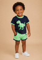 A smiling toddler stands against a warm beige studio background wearing a navy blue short-sleeve T-shirt with green gingham trim and a green dinosaur graphic, matching green gingham shorts, and white sneakers.
