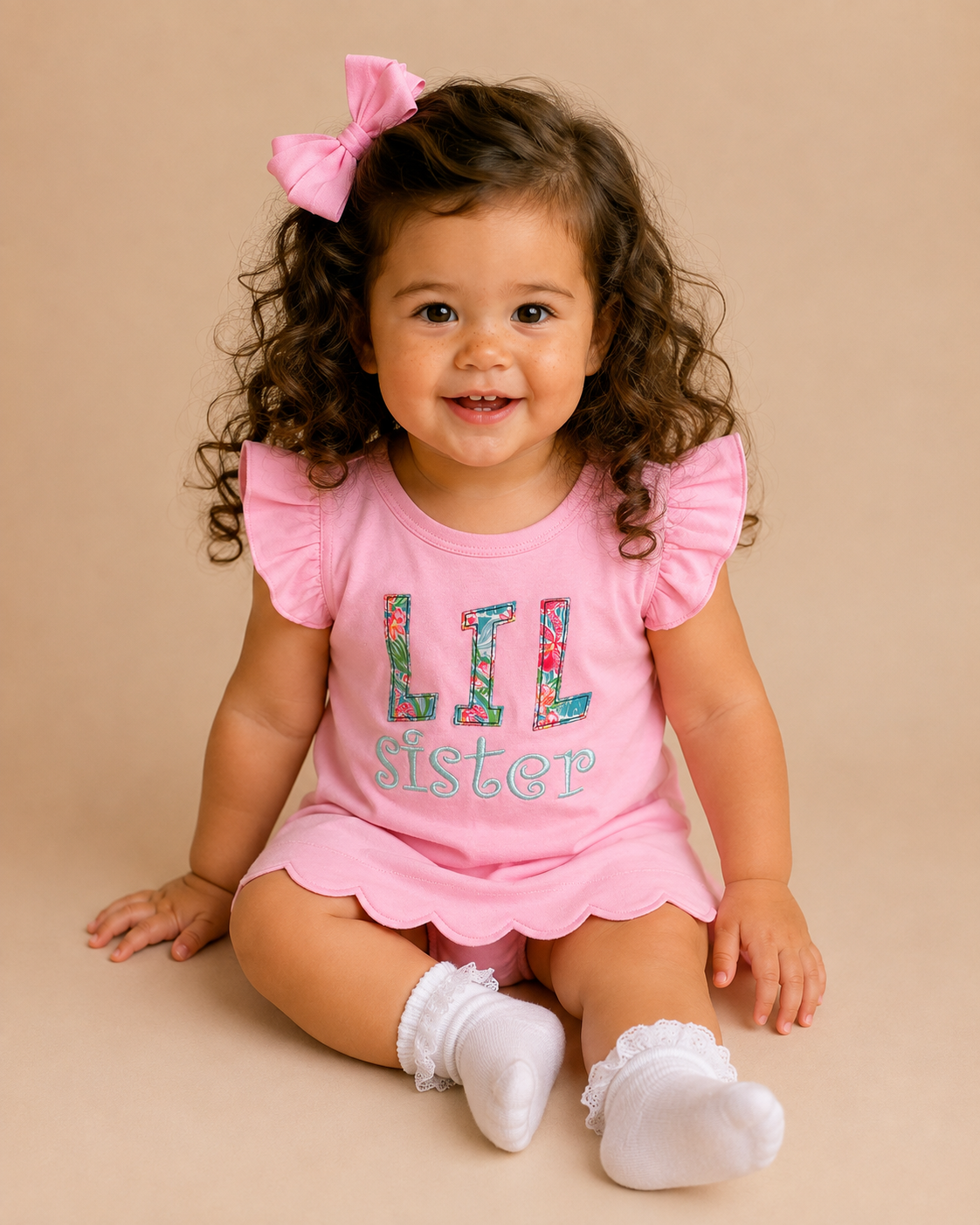 Smiling toddler with LIL SISTER embroidered on the front of a pink, scalloped hem shirt.