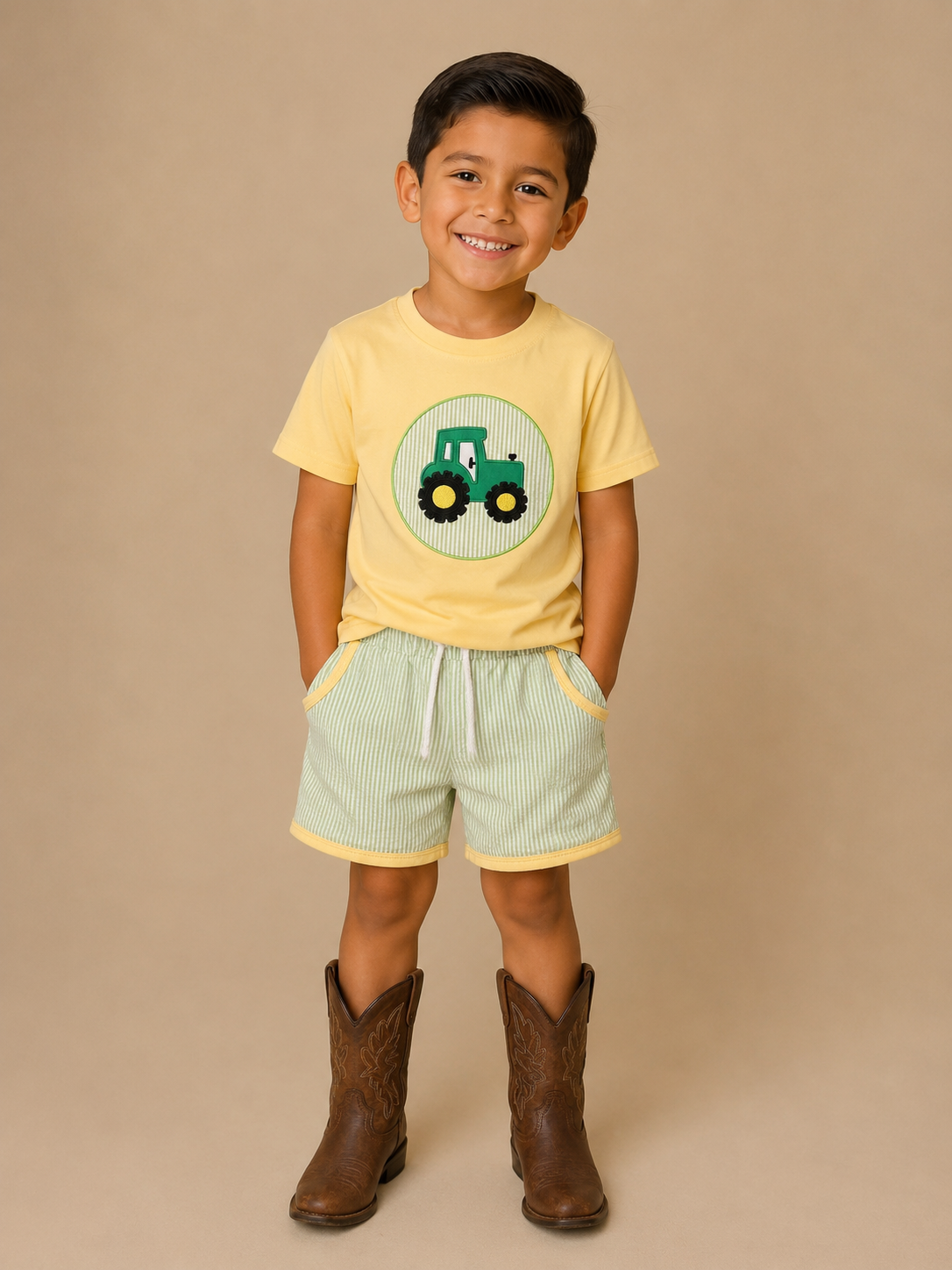 Child wearing a yellow t-shirt with a green tractor design, light green shorts, and brown cowboy boots on a beige background.