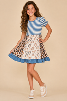 A young girl modeling a short-sleeve tiered dress with a blue and white striped bodice and a rainbow-and-leopard-print skirt, posed against a solid beige studio background.