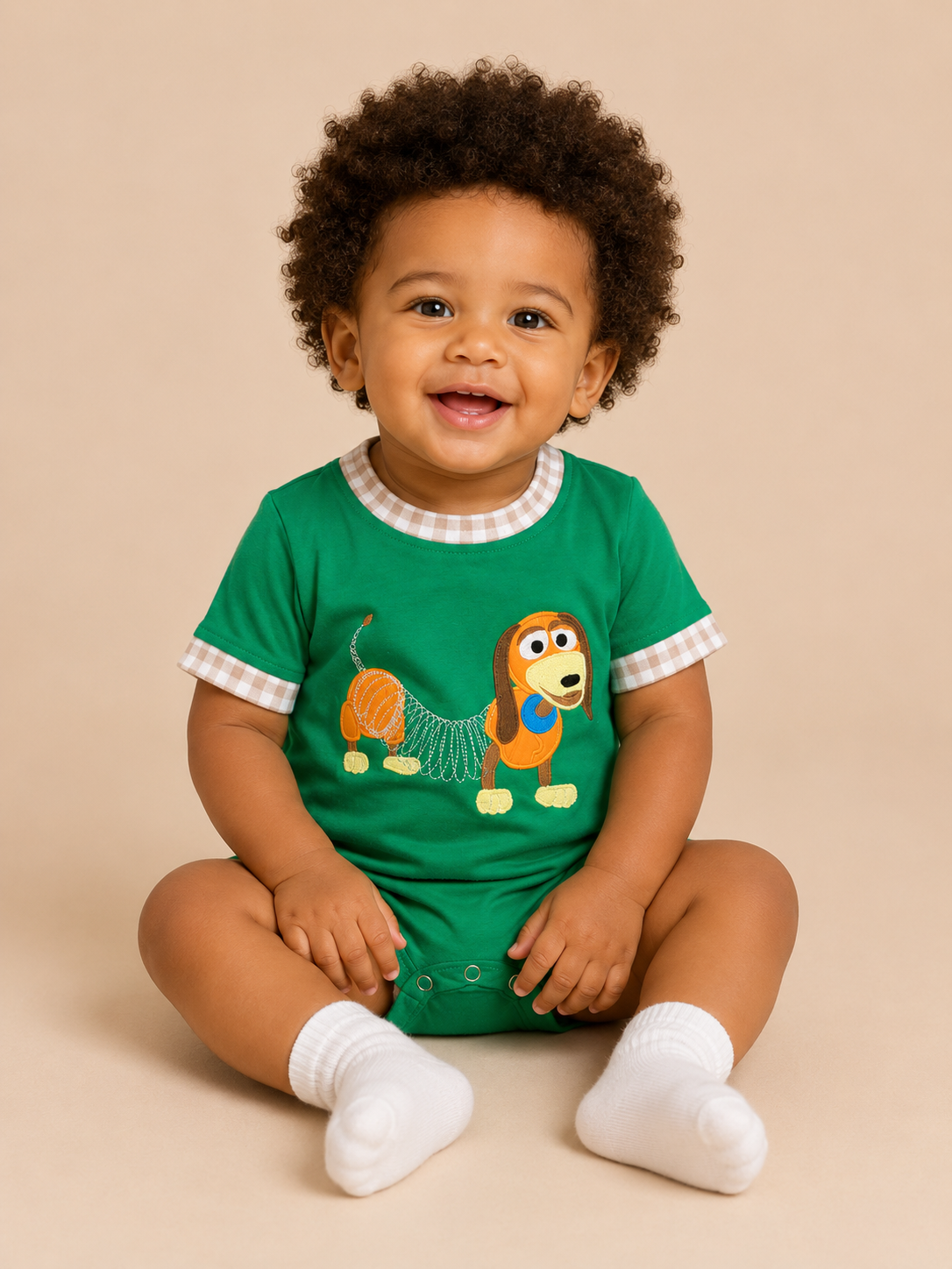 A smiling baby with curly dark hair sits cross-legged against a solid beige background, wearing a green short-sleeve bodysuit with a cartoon dog graphic and white socks.