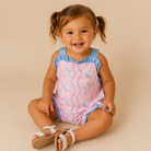 A smiling toddler girl with brown hair in pigtails sits cross-legged on a warm beige studio background. She is wearing a sleeveless pink romper with blue ruffled trim and a seashell and starfish print, along with white sandals.
