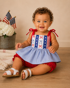 Baby wearing a red, white, and blue dress with bow accents on a wooden floor.