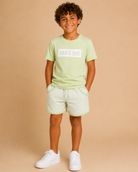 Child wearing a light green t-shirt with 'DAD'S BOY' text and matching shorts on a beige background