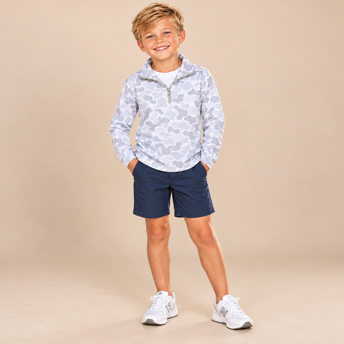 A young boy stands against a solid beige studio background wearing a light gray and white camouflage patterned quarter-zip pullover, navy blue shorts, and white sneakers.
