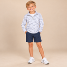 A young boy stands against a solid beige studio background wearing a light gray and white camouflage patterned quarter-zip pullover, navy blue shorts, and white sneakers.