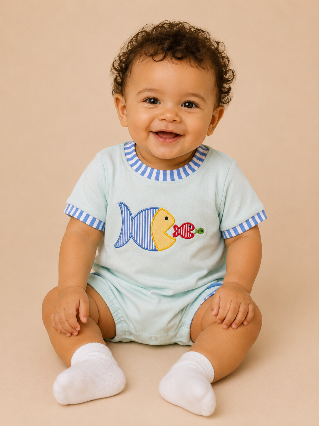 A smiling baby with curly dark hair sits on a beige studio background wearing a light blue short-sleeve bubble with a striped blue-and-white collar and sleeve cuffs, plus a fish graphic on the chest. The baby is also wearing white socks.