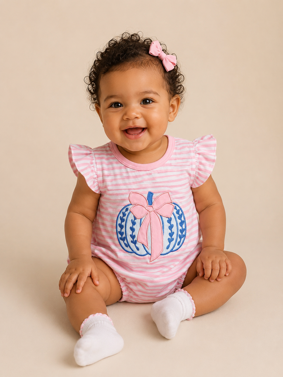 A smiling baby girl with curly hair and a pink bow sits on a beige studio background, wearing a pink and white striped short-sleeve romper with a blue and pink pumpkin graphic on the front, along with white socks.