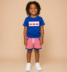 A young child with dreadlocks stands against a solid beige background wearing a royal blue t-shirt with a red and white train graphic, red and white striped shorts, and white sneakers.