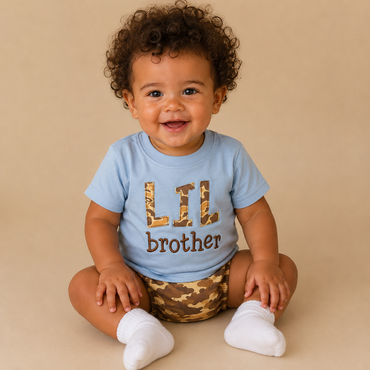 A smiling baby with curly dark hair sits cross-legged on a beige studio background, wearing a light blue T-shirt with 'LIL brother' printed on the front in leopard-print and brown lettering, plus camouflage-patterned shorts and white socks.