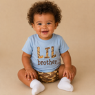 A smiling baby with curly dark hair sits cross-legged on a beige studio background, wearing a light blue T-shirt with 'LIL brother' printed on the front in leopard-print and brown lettering, plus camouflage-patterned shorts and white socks.