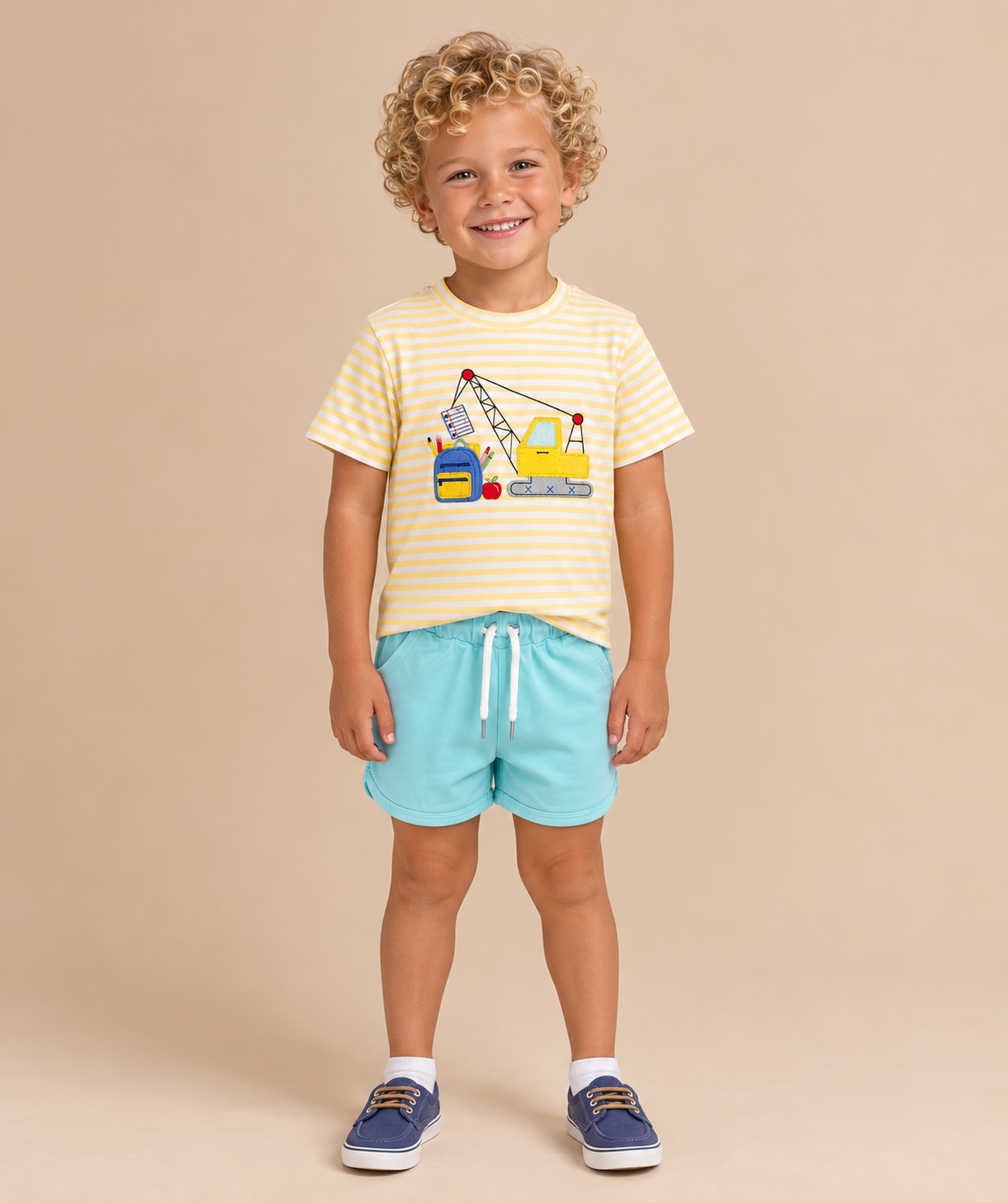 A young child with curly blonde hair stands against a solid beige studio background, wearing a yellow and white striped t-shirt with a construction crane graphic, light blue shorts, and navy blue shoes.
