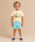 A young child with curly blonde hair stands against a solid beige studio background, wearing a yellow and white striped t-shirt with a construction crane graphic, light blue shorts, and navy blue shoes.