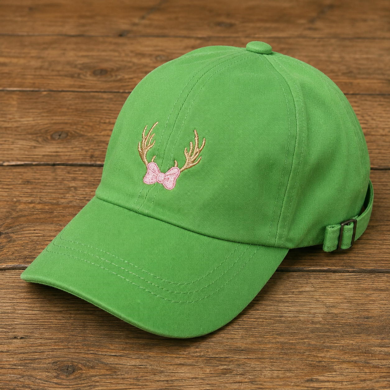 A green baseball cap with an embroidered pink bow and gold antlers on the front, displayed on a rustic wooden surface.