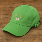 A green baseball cap with an embroidered pink bow and gold antlers on the front, displayed on a rustic wooden surface.
