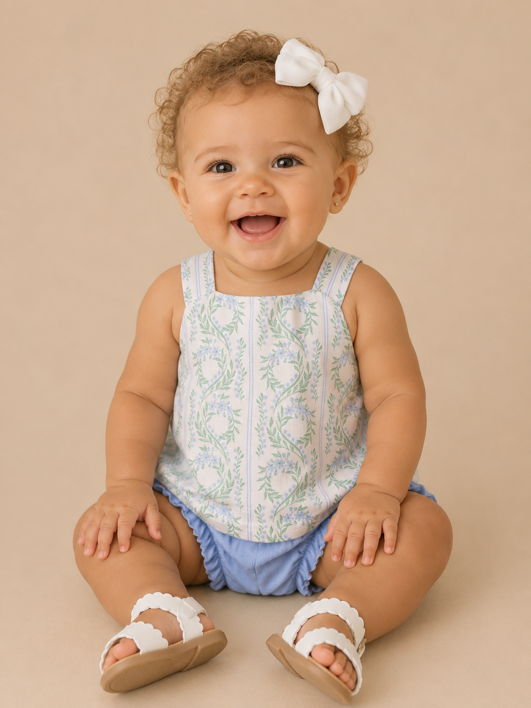 Baby sitting on a beige surface wearing a floral top and blue shorts with white sandals.