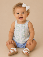 Baby sitting on a beige surface wearing a floral top and blue shorts with white sandals.