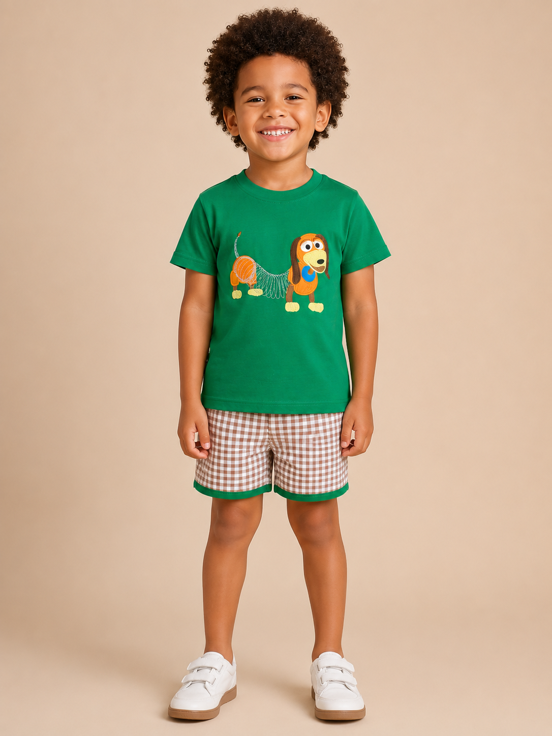 A young boy stands against a solid beige studio background wearing a green short-sleeve t-shirt with a cartoon dog graphic, brown and white gingham shorts with green trim, and white sneakers.