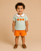 A toddler standing against a solid beige studio background, wearing a short-sleeve polo shirt with green and white horizontal stripes, three orange pumpkins on the chest, a white collar, and bright orange shorts.