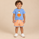 Toddler boy wearing a light blue shirt with a pumpkin goose wearing orange and white gingham shorts.