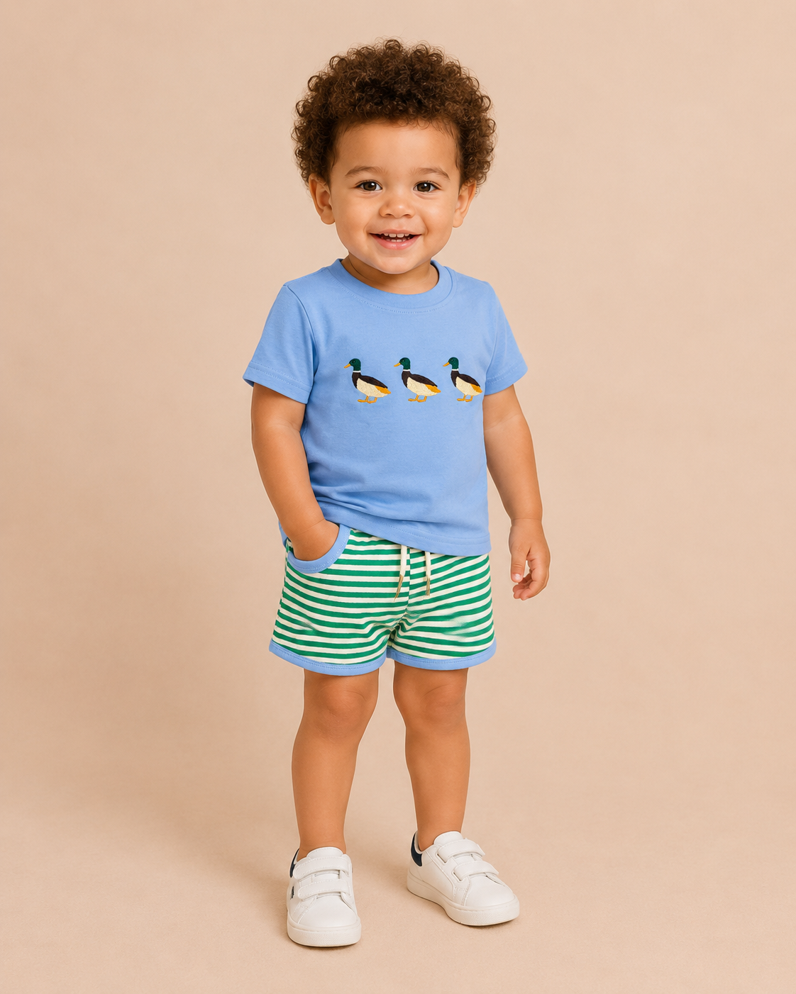 Toddler boy wearing a light blue shirt with three ducks on the front with green striped shorts.