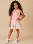A young girl with long curly brown hair stands against a solid beige studio background, wearing a pink sleeveless polo shirt with a dog patch on the chest, a pink and white gingham pleated skirt, and white sneakers.