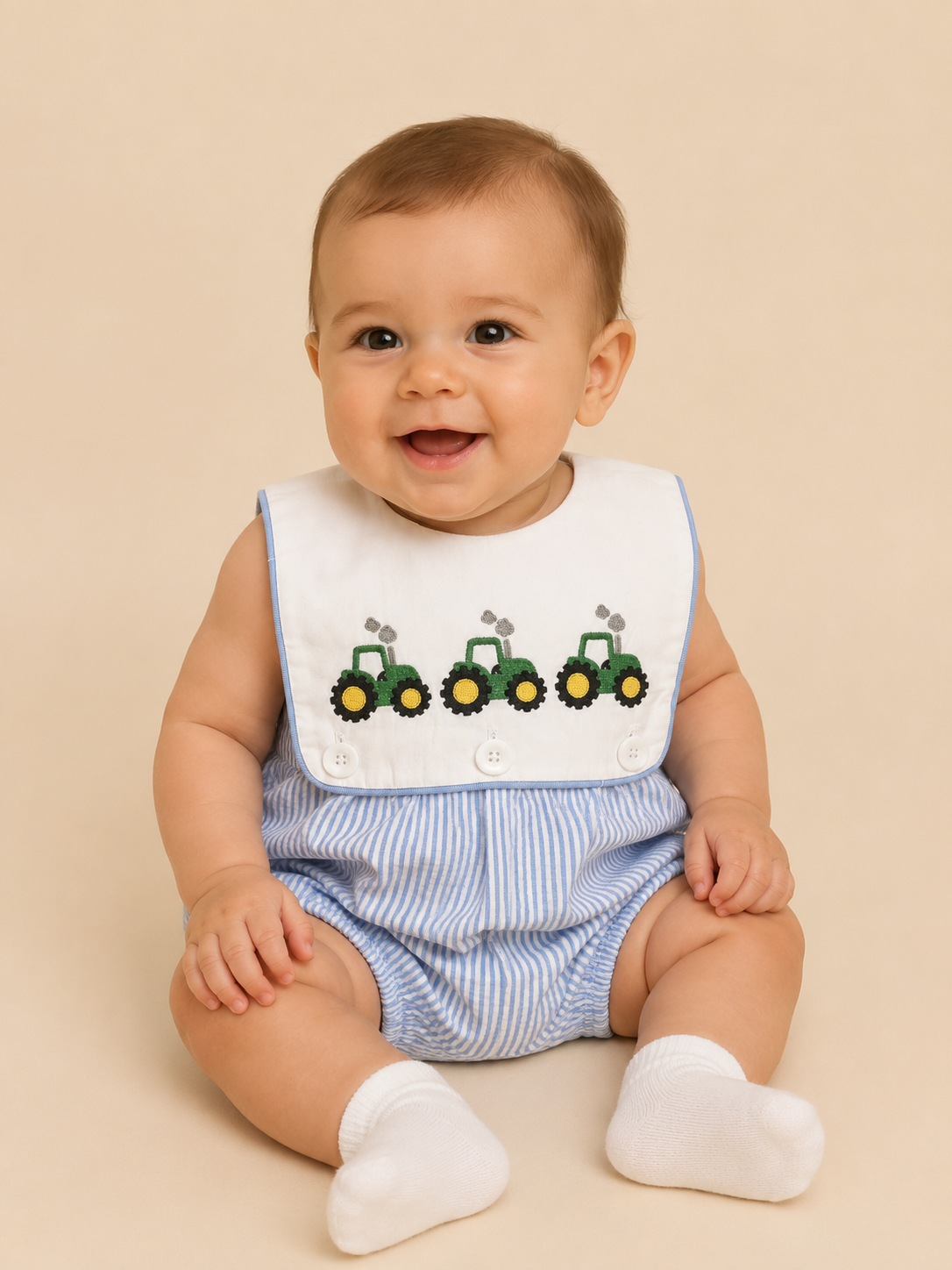 A baby wearing a white bib with three green tractors and blue-and-white striped romper, sitting against a solid beige background.
