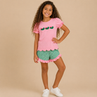 Young girl wearing a pink shirt with French knot alligators, scalloped hem, gingham green shorts.