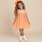 A young girl with curly brown hair wearing an orange and white gingham dress with a white Peter Pan collar decorated with three orange pumpkin appliques. She is also wearing an orange bow headband, white socks, and white shoes, standing against a solid beige studio background.