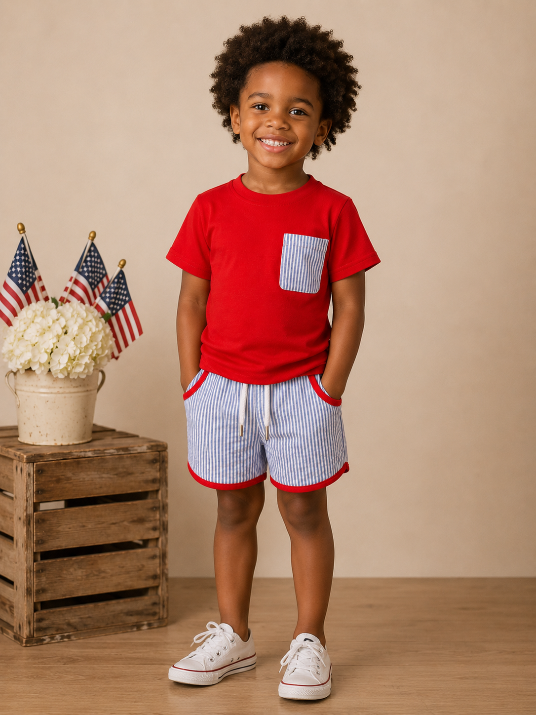 Child wearing a red shirt and striped shorts standing in front of a wooden crate with flowers and American flags.