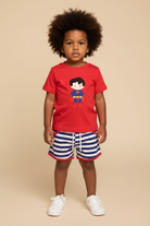 Toddlerboy with afro wearing red graphic embroidered tee and blue white striped shorts with white sneakers on beige background