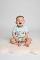 A baby sitting on a white faux fur rug wearing a mint green short-sleeve bodysuit with a yellow and blue fish graphic on the front, set against a plain light gray studio background.