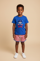Young boy wearing a blue t-shirt with a super hero embroidery, red and white striped shorts, beige background.