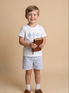 Child wearing a white t-shirt with blue crosses, shorts, and brown shoes, holding a book on a beige background