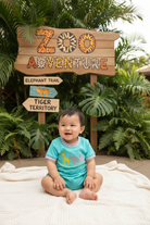 Child in a turquoise outfit with animal prints sitting on a blanket in front of a 'Zoo Adventure' sign.